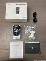 Full view of the camera, mounting bracket with hardware, charging cable, user manual, warranty card, and box arranged on a flat surface.