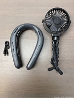 Image showing black flexible mini fan with adjustable tripod legs and gray U-shaped neck fan with USB cable, laid flat on beige surface.