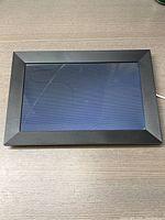 Black digital picture frame on a wooden surface, showing the screen turned on with some reflections and smudges visible.