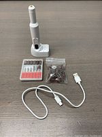 Electric nail file set displayed on a wooden surface including the main device in its stand, filing attachments, sanding bands, and charging cable.