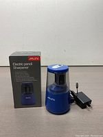 Electric pencil sharpener with its box and the power adapter laid out on a neutral background.