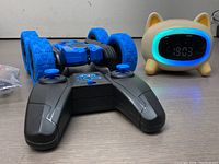 Rear angled view of blue remote control stunt car with dual joystick controller, alongside beige cat-shaped LED clock with glowing blue LED frame around digital display.