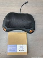 Black electric handheld massager with mesh surface and orange control buttons, placed beside a labeled boxed gel bead face mask.