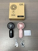 Two mini fans in black and pink with packaging, USB cables, and instructions on a wooden surface.