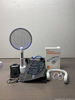Lot display of four outdoor items including mosquito zapper, neck fan, backpack, and insect swatter on neutral background