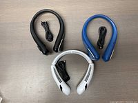 Top-down photo showing all three neck fans in black, white, and blue with their USB charging cables, arranged on a tabletop.