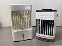 Two portable air cooler fans side by side showing front and top controls, white compact designs with vents and control buttons.