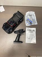 Overall view showing black RC car, remote control, spare parts in sealed packet, and user manual on a wooden table.