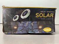 Box showing product packaging labeled 'Solar Powered Solar Disk Lights' 12 pack with images of round disk lights and lit pathway.