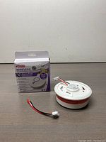 Photo showing the Kidde wireless smoke and carbon monoxide alarm device along with its original retail packaging and an additional wiring connector laying on a wooden surface.