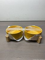 Front view showing two yellow deep circular dog bowls side-by-side, each secured with clear tape and white liners. Bowls rest on a wooden stand with block supports.