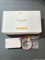 Photo of the white packaging box labeled Luminoodle Click Stick-Anywhere Lighting 3-Pack with one LED light strip coiled and connected to a white battery pack with a touch button, along with the product manual.