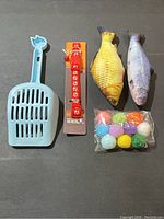 Photo of all items in the cat essentials bundle displayed on a flat surface including blue litter scoop, red bell collar, two fish toys, and pom pom balls
