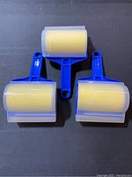 Set of 3 blue plastic pet hair lint rollers with pale yellow adhesive sheets and protective covers arranged on a dark surface