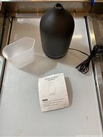 Black conical essential oil diffuser, power cord, clear plastic measuring cup, user manual on metal surface