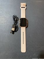 Full view of smart watch with beige strap and USB charging cable.