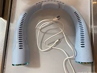 White electric neck fan with a curved U-shape and ventilation slits along the sides, lying flat on a surface with included USB charging cable.