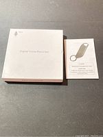 White box labeled Digital Voice Recorder and user manual placed side by side on a dark surface.