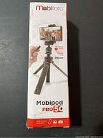 Front view of the Mobifoto Mobipod Pro 50 tripod box showing the tripod with smartphone attached and Bluetooth remote shutter.