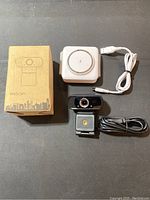 Wired USB webcam with clip and black USB cable beside white wireless charging docking station with USB cable, original webcam box included.