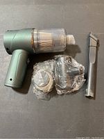 Mini handheld vacuum cleaner in metallic green with transparent dust container alongside three cleaning attachments, two wrapped in plastic.
