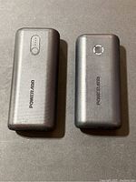 Two silver Poweradd 10,000 mAh power banks side by side on a brown surface, showing top view with different button designs.
