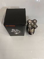 Photo showing the Maxgroom 5D Head Shaver next to its original box, featuring the shaver with five rotary blades arranged in a cross pattern and ergonomic handle.