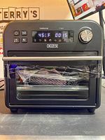 Front view of CKOZESE air fryer oven showing digital touchscreen display, door with glass window, stainless steel handle, and visible trays inside wrapped in plastic.