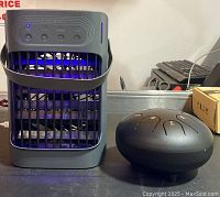 Photo showing the portable cooling fan with front grill, control buttons, blue LED lights, and handle, next to the black steel tongue drum with tongue slits.