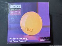 Box front showing Dekala brand, product image of round sunrise alarm clock, and key features including 2-year warranty and 30-day money-back guarantee.