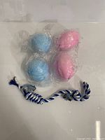 Four rubber chew balls, two blue and two pink, individually wrapped in plastic, arranged on a surface with one blue and white braided rope chew toy.