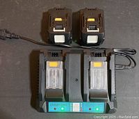Two black lithium-ion drill batteries resting on a black dual charging dock with green and red indicator lights. The dock model CHAMT-2042 is visible and the dock is powered on as indicated by the green light.