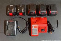 Five lithium-ion drill batteries and two charging docks laid out on a gray surface. Batteries show red and black color accents, chargers have attached power cords.