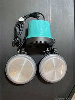 Electric water pump with teal top housing and black base, power cord attached, shown next to two circular wall lights.