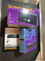 Image showing three boxed media players: Proscan DVD player with progressive scan, Sylvania HDMI DVD player, and a Bluetooth CD micro system.