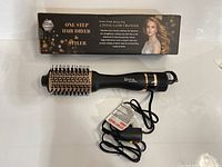 Black one step hair dryer and styler brush with ceramic-coated bristles next to its box, showing the product and power cord with tag.