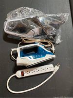 Blue and white steam iron, white power strip with multiple outlets, black and white handheld vacuum in clear plastic bag, arranged on dark surface.
