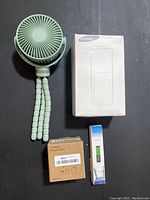 Top-down view of all four items on black surface: mini fan with flexible legs, boxed soap dispenser, small timer box, water tester packaging.