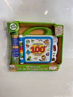 Front view of the LeapFrog 100 Words Book in original packaging with visible orange handle, blue stylus, and colorful cover.