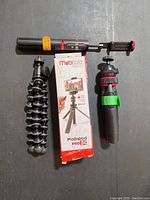 Photo of two mini handheld tripods and two selfie sticks with the Mobipod Pro 5G packaging box on a dark surface