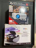 Boxes of the RCA home theater projector and Proscan radio cassette CD boombox stacked with visible branding and features.