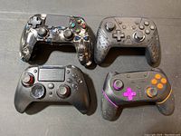 Four various gaming controllers displayed top-down, showing front face including buttons, joysticks and directional pads. Controllers show wear and use signs.