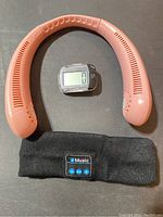 Top view of pink neck fan, black headband speaker featuring music control buttons, and clip-on pedometer with digital display on black surface.
