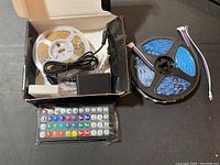 Photo showing two rolls of LED strip lights, one with white backing and the other with black backing, power adapter, and remote control laid out on a dark surface inside a box.