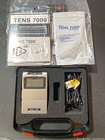 Top down view of TENS 7000 device, electrode cables, user manual, electrode pads packet in foam case interior.