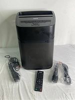 Front view of the black portable projector with ventilation and speaker grille. Two wireless microphones and remote control are displayed beside it, all items appear in good condition.