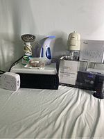 Full view of all items in the lot including garment steamers, power bars, USB hub, Bluetooth CD micro system, baby food maker on its box and some accessories.