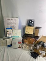 Various items including cooling fan, video switch, earbuds, cables, light bulbs, and other tech and home items on a bed surface.