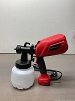 Side profile of red and black electric paint sprayer with white plastic container and black cord lying on surface