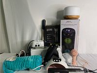 Photo showing multiple personal care devices including hair clippers, massage tool, nail grinder, UV nail lamp, heating pad, hot comb, hair straightener, Bluetooth speaker, essential oil diffuser, and mini fan arranged on a surface.